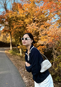 A woman wearing sunglasses leaning forward beside a road lined by trees on a sunny autumn day.