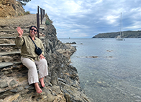 A woman sitting on stone steps smiling and waving beside a body of water on a sunny day.