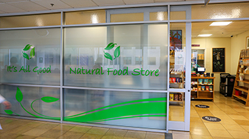 A worker wearing plastic gloves uses tongs to pick up grilled vegetables while holding a partly filled takeout container.