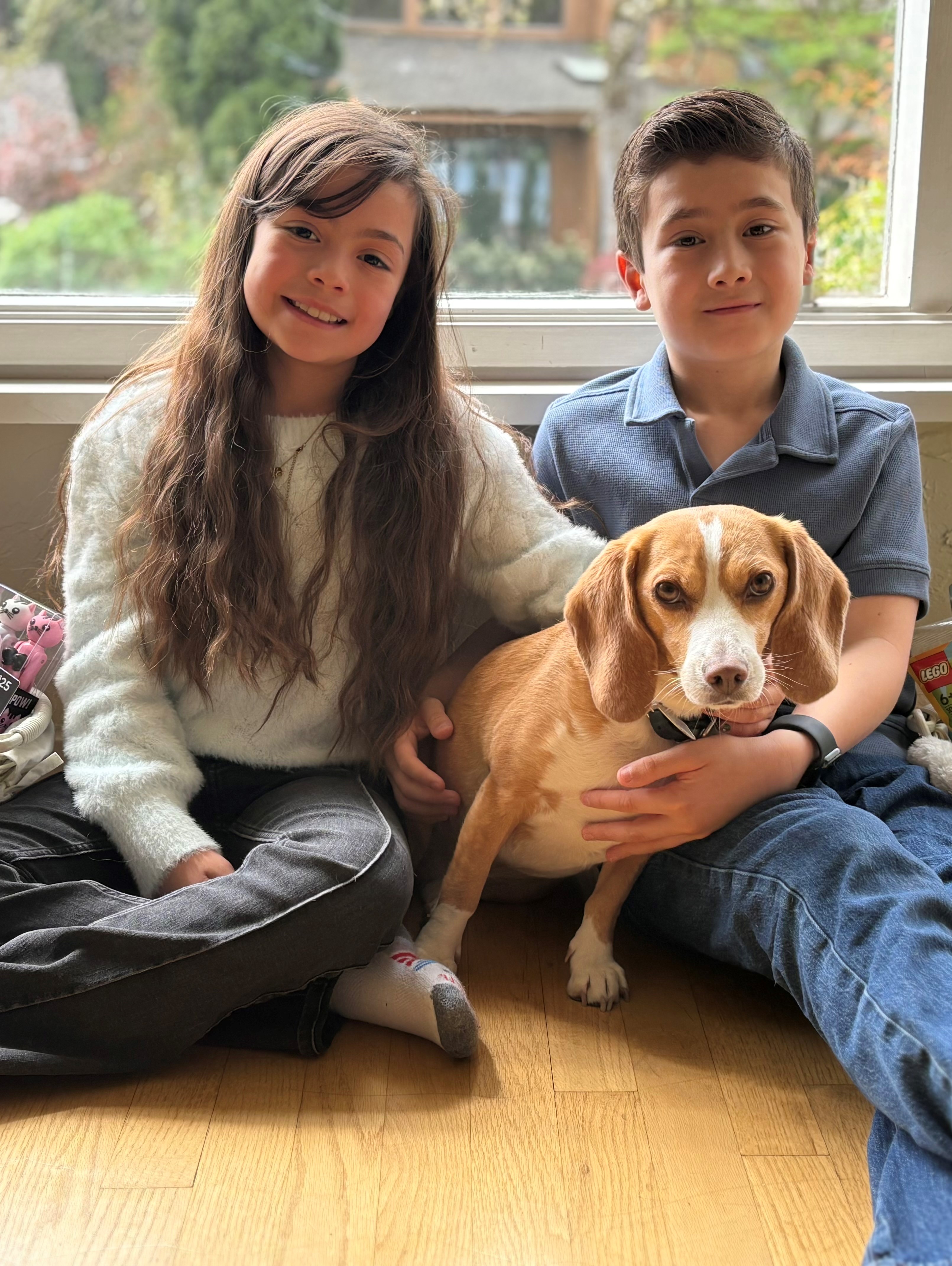 Two kids sit side by side smiling at the camera with a dog sitting between them.