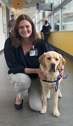 A smiling woman with shoulder-length brown hair, dressed in a black top and khaki pants, crouches next to a standing dog, her hands on the dog’s shoulders. The dog’s leash is entwined in her left hand.