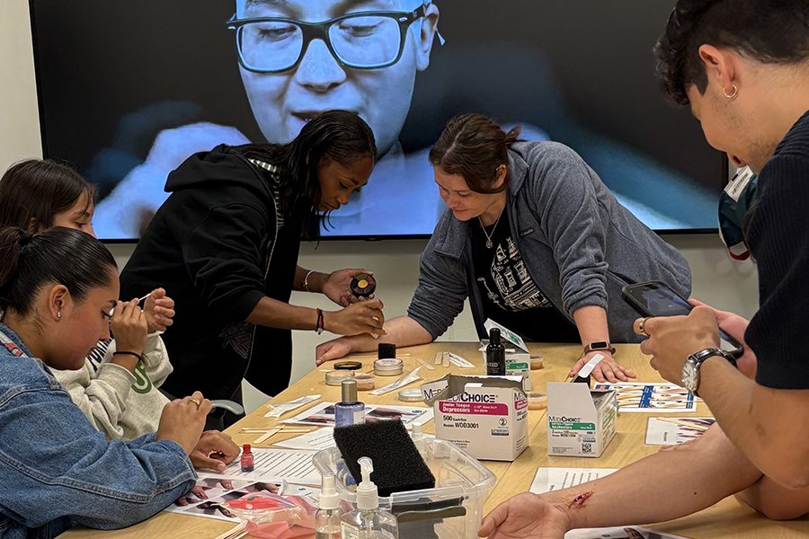 A group of six people around a table practicing wound treatment as a demonstration video plays on a TV behind them.