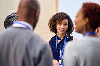 A small group of faculty and educators in business attire gather during a workplace meeting or discussion. The focus is on a woman with curly hair speaking to the group.