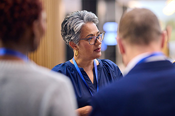 Educators engaged in collaborative discussion. A woman in glasses and navy blue attire speaks with colleagues in a professional setting.