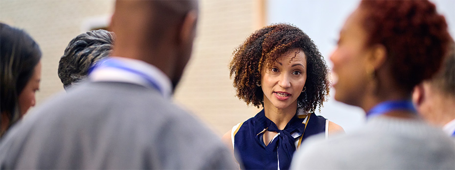  A small group of faculty and educators in business attire gather during a workplace meeting or discussion. The focus is on a woman with curly hair speaking to the group.