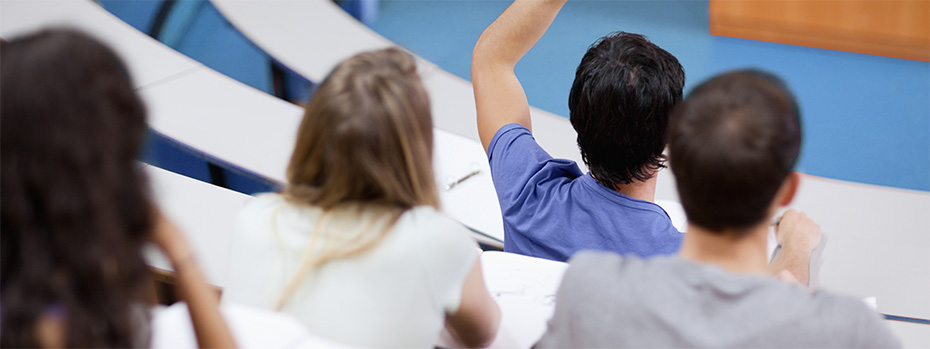 OHSU students sit in a classroom with hands raised, viewed from behind. One student in a blue shirt has their hand up to ask or answer a question.
