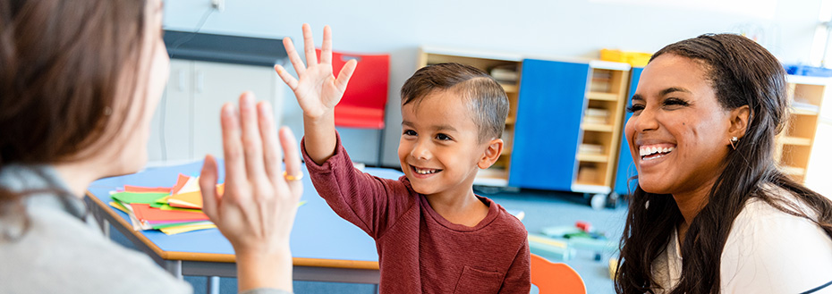 Teacher gives preschool-age boy a high five across the table in a classroom while his mom looks on smiling.