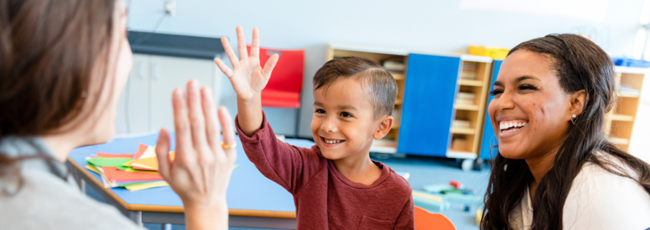 Una maestra choca los cinco con un niño en edad preescolar sentado al otro lado de la mesa en un aula, mientras su madre observa sonriendo.