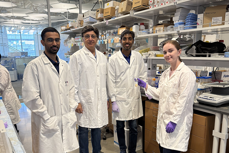 Four researchers wearing white coats standing in a laboratory. Three of them are men and one is a woman, with the woman holding a test tube.