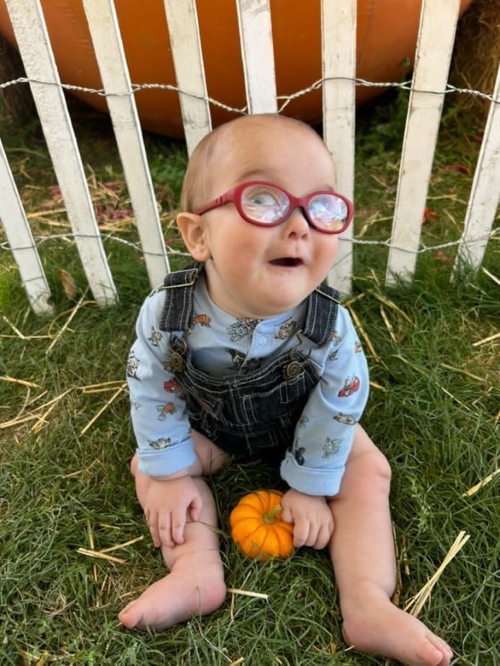 A baby sits in the grass holding a mini pumpkin looking up at the camera wearing red glasses.