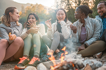 Students sit and socialize around a campfire against the backdrop of mountains and forest.