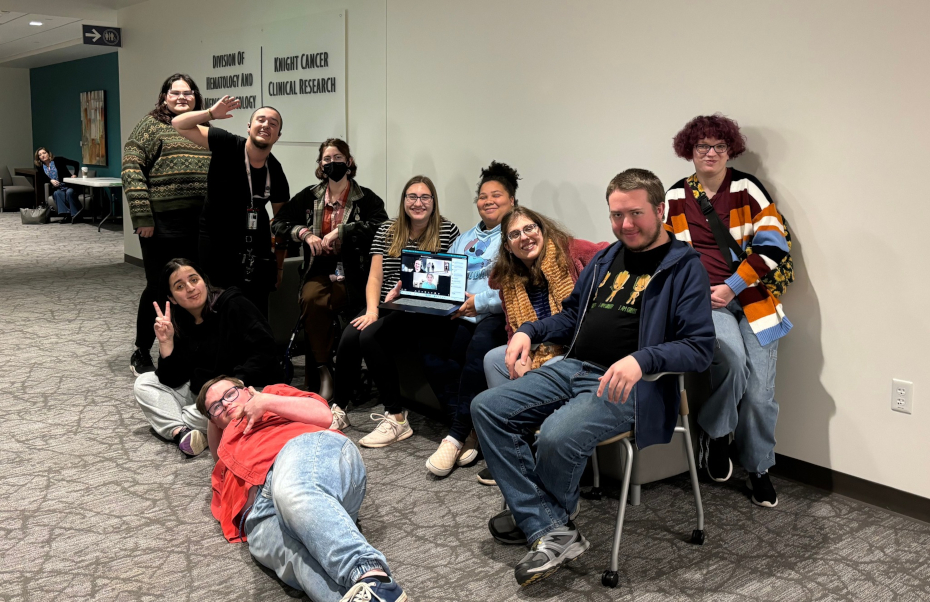 Ten of the youth advisory board members smile and pose together at the Fall 2024 retreat. One of the members holds a laptop showing some of the members that attended virtually on a video call on the screen.