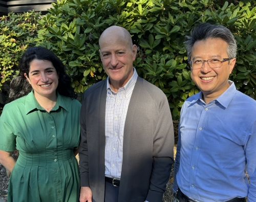 Two men and a woman stand shoulder to shoulder smiling at the camera in front of a green ivy background.