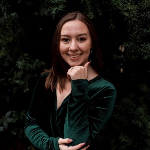 Ashley, a woman with brown hair and a dark green dress stands in front of dark pine trees. She is smiling and has one hand under her chin.