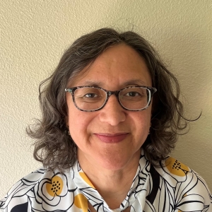 Cristela, a woman with dark, curly hair and glasses smiles at the camera, wearing a white shirt with a black and yellow floral pattern.