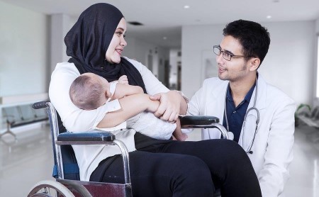 A hijabi woman is holding a baby while seated in a wheelchair, a doctor is kneeling beside her to talk at eye level