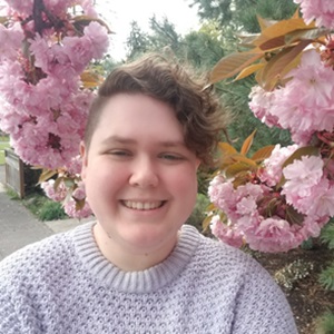 Jay, a white person in their 30s, wearing a lavender sweater is smiling in front of two branches of blooming cherry tree pink flowers