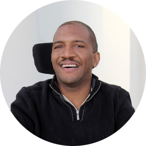 Dr. Lateef McLeod. A Black man with a buzz cut smiles as he sits at a desk in a power wheelchair. He is wearing a black quarter-zip sweater.