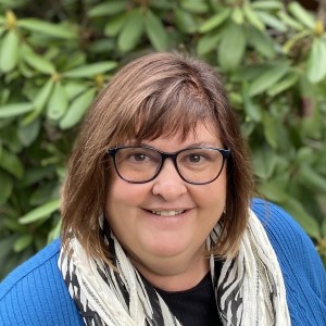Rhona, a middle-aged woman with shoulder length brown hair, wearing round glasses and a scarf, is standing in front of greenery and smiling