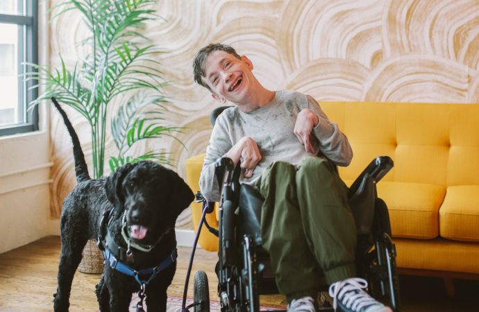 Sex and Disability Educator Evan Sweeney is a 42-year-old disabled male with light skin and short brown hair wearing a gray long-sleeve shirt, forest green pants and navy blue Converse shoes. He sits in a power wheelchair smiling brightly next to his black medium size service dog.