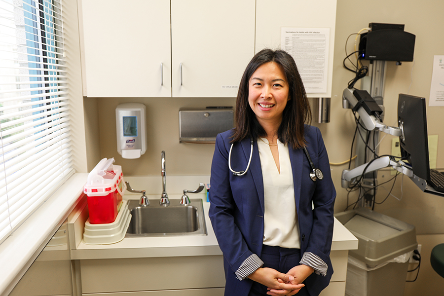 A female doctor stands next to the window in an exam room with a stethoscope around her neck.
