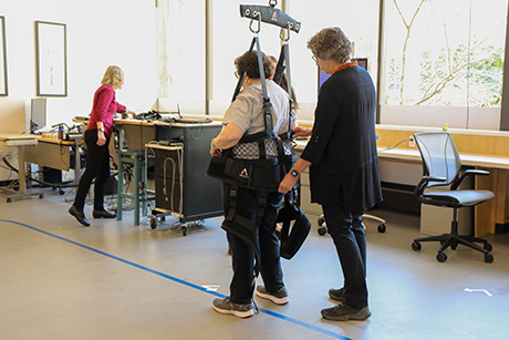 A patient in a harness attached overhead is guided by staff members as she prepares to follow a path marked in blue tape. 