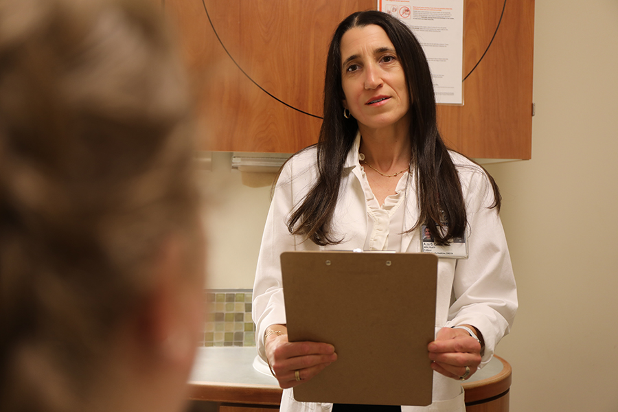 A woman standing with a white physician coat on holding a clipboard speaks to a patient in an exam room.