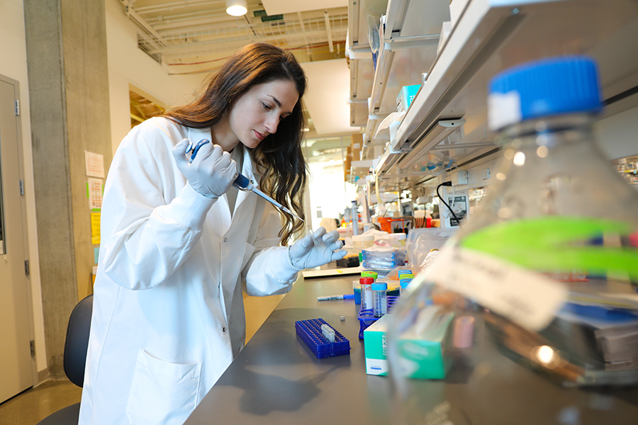 A female researcher in a research lab wearing a white lab coat fills a test tube with a liquid sample.