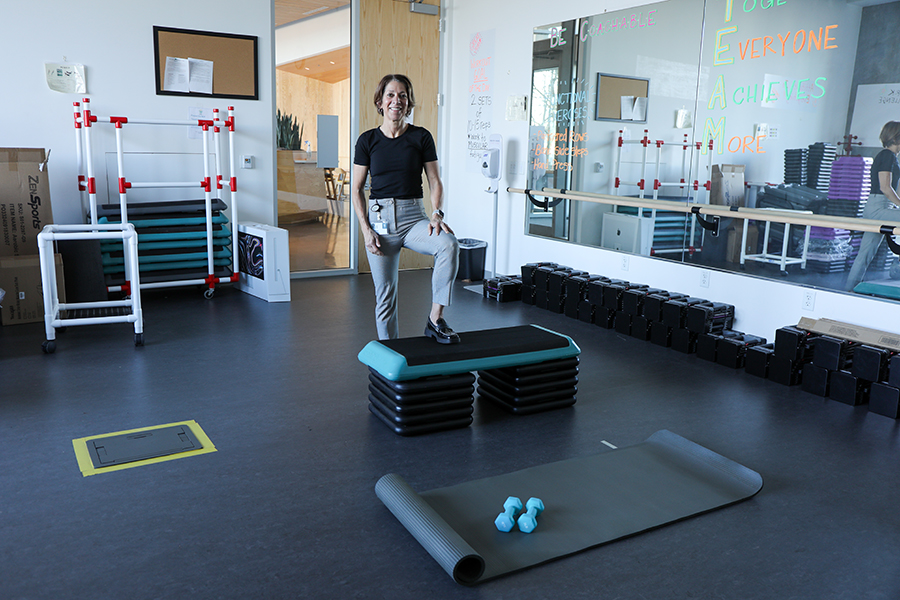 A woman stands smiling with one leg up on a step in a rehab room.