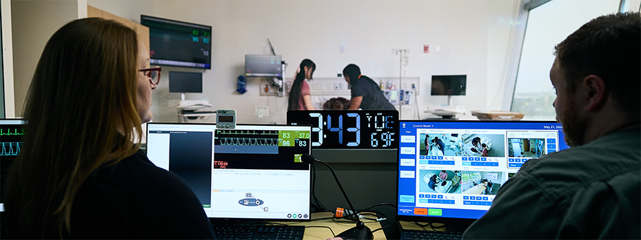 Two OHSU students monitor vital signs on computer    screens and observe simulations from the control room during    nursing training.