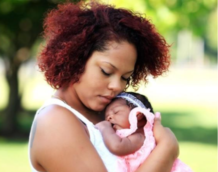 A woman wearing a white shirt holds a baby in a pink dress wearing a white headband.