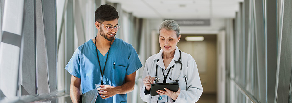 A young male doctor in scrubs and an older female doctor in a white doctor coat talk as they walk down a well-lit hallway with windows and steel railing.