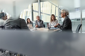 A group of health care and business professionals engaged in a leadership discussion around a conference table in a meeting room.