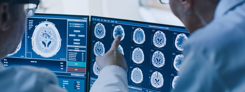 Two men in white lab coats look at two computer monitors showing multiple brain scans as one person point at one of those images