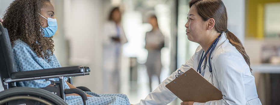 Female doctor in a white coat kneels to comfort a girl in a wheelchair wearing a mask.