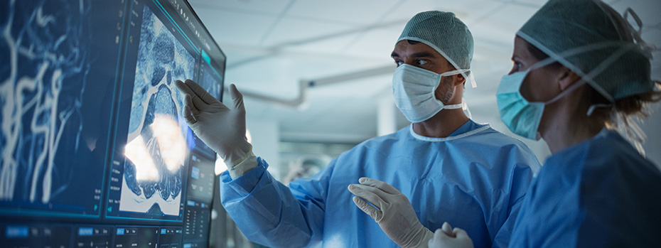 Two doctors in scrubs and masks review digital scans on a large screen in a dimly lit operating room. 