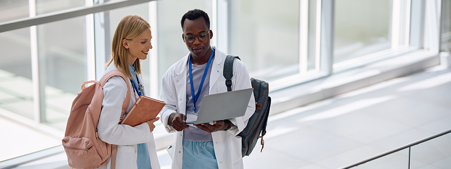 A young female doctor and male doctor stand in an empty hallway lined with windows reading a laptop screen