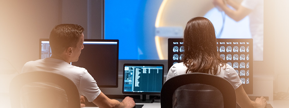 Two medical physics students reviewing scans on multiple monitors in a radiology reading room.