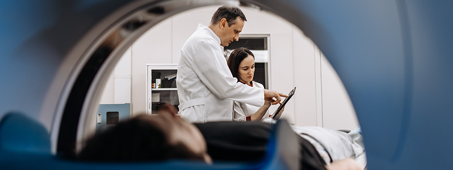 Two medical physicists review patient imaging data on a tablet in front of a CT scanner.