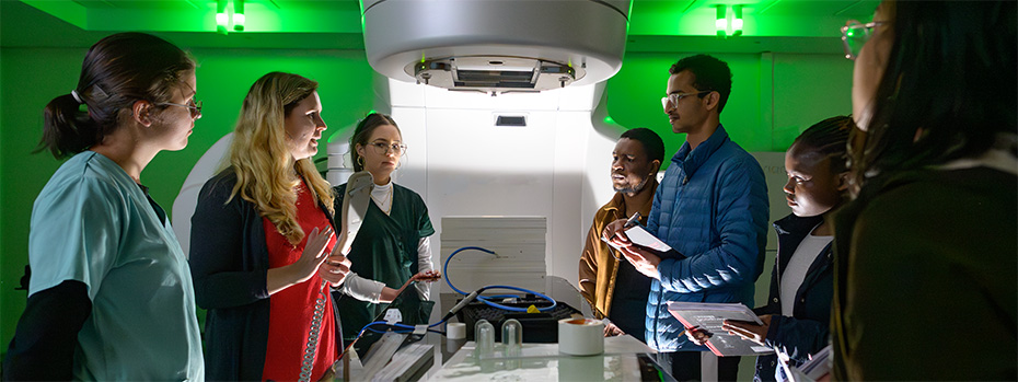 A group of medical physics students and professionals gathered around a linear accelerator during a training session.