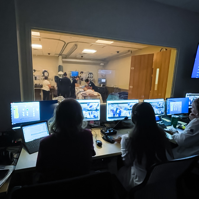 Simulation technicians monitor a live training scenario from a control room at Sky Lakes Collaborative Health Center in Klamath Falls, observing OHSU students through an observation window and multiple screens.