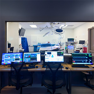 A control room in the Mark Richardson Interprofessional Simulation Center at OHSU, with multiple monitors displaying patient vitals and simulation software, overlooking a simulated operating room through an observation window.