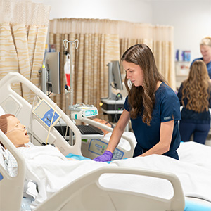 An OHSU nursing student in scrubs performs a bedside care skill on a patient manikin in a simulation lab.