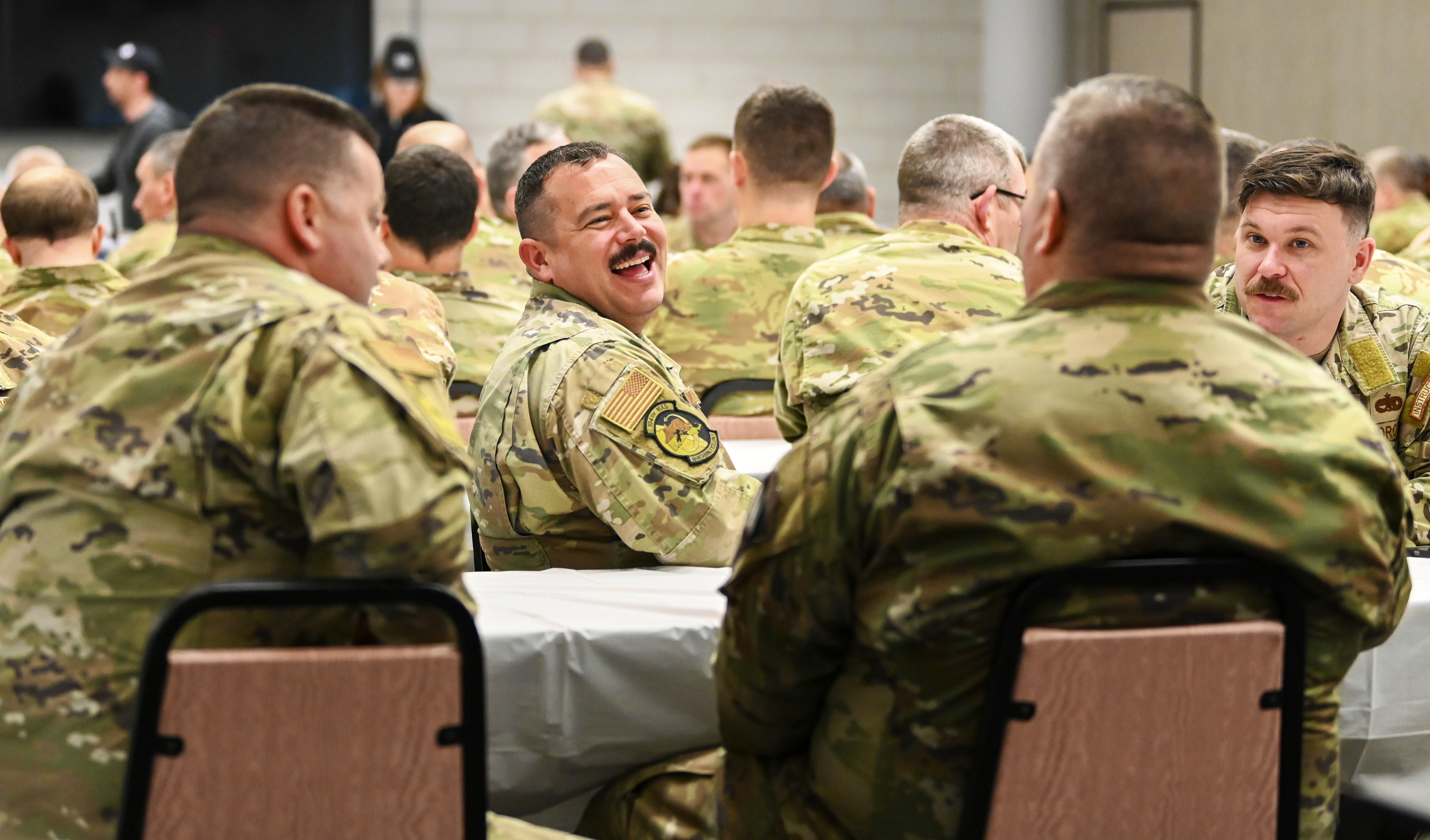 A group of U.S. military service members in camouflage uniforms sit together at tables in a large indoor venue, engaged in conversation. At the center of the frame, one service member laughs broadly while talking with colleagues. Additional service members fill the background, creating an atmosphere of camaraderie and connection.
