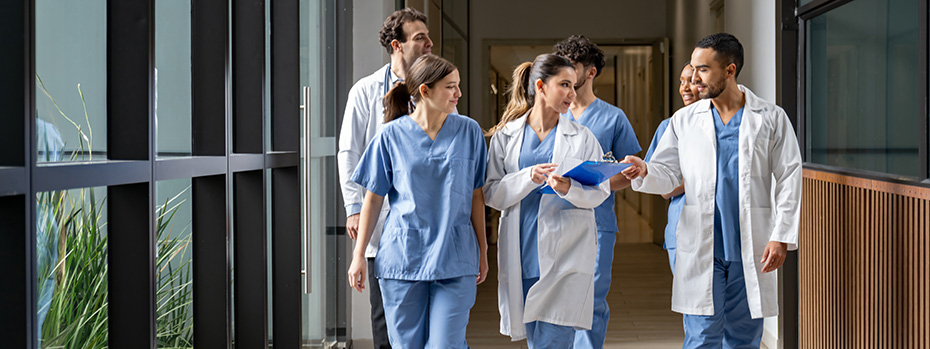 Six young doctors—three in white coats and three in blue scrubs—talk while walking down a wide well-lit indoor corridor framed by a wall of windows
