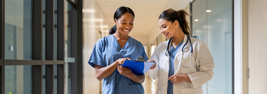 Two young female doctors smile and talk as they walk through a hall lined with windows.