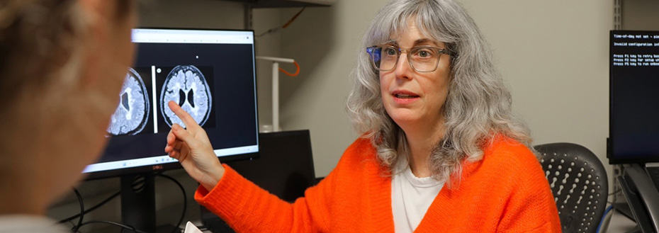 Dr. Rebecca Spain confers with a colleague. She is seated at a desk and points to brain images on a monitor while looking at her colleague.