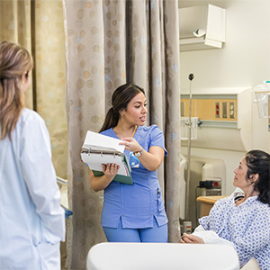 A OHSU nursing student reviews patient information with a standardized patient in a simulated hospital room while a clinical instructor observes.