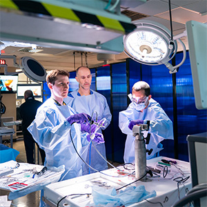 Three OHSU students in surgical gowns and gloves perform a hands-on tissue-based simulation using instruments and equipment in an operating room–style lab.
