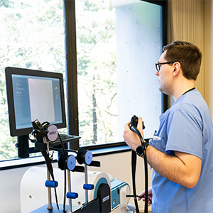 An OHSU student in scrubs practices with a virtual reality endoscopy simulator while viewing a monitor.